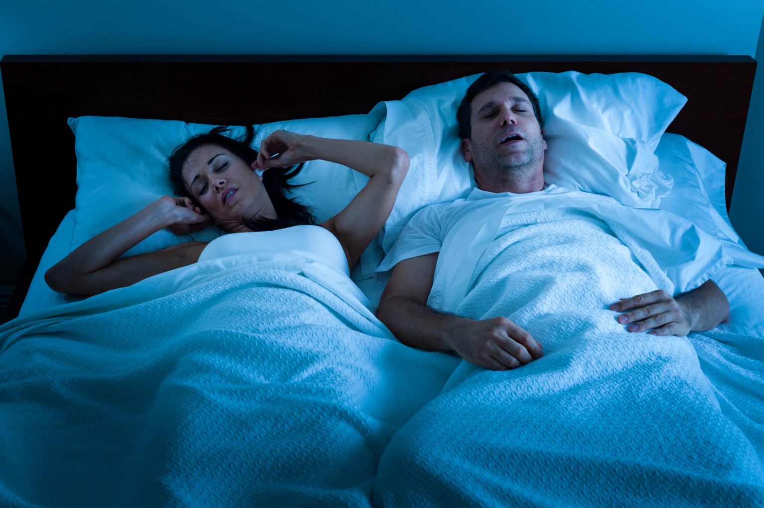 Man and woman in bed and the woman is covering her ears due to the man snoring.